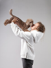 happy girl with a dog in a photo studio on gray. Curly woman in a white hoodie with a chocolate poodle in her arms. - obrazy, fototapety, plakaty