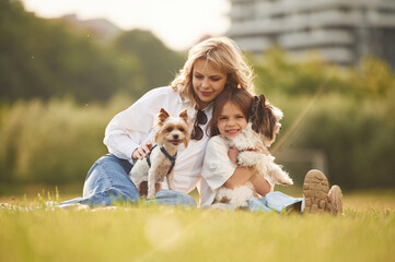 Naklejka na ściany i meble Sitting together. Beautiful mother with her little daughter and dogs are on the summer field