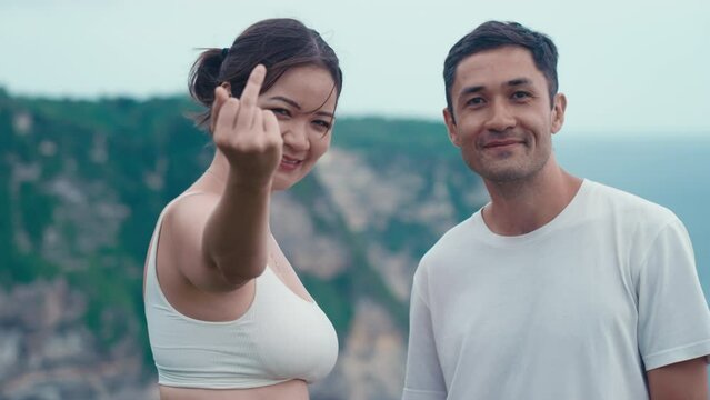 Asian people posing for photograph. Sneaky girl with mean smile showing obscene gesture to camera to make her boyfriend laugh. Korean travelers. Vacation on a rocky ocean shore. Rebel gesture.