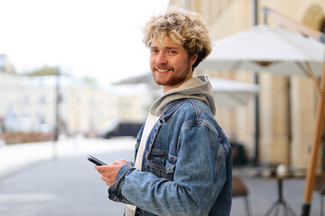 A young guy in casual clothes holds a mobile phone in his hands and smiles against blurred street...
