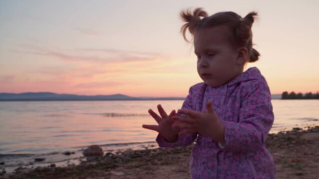 A little girl on the shore of the lake at sunset collects sand in her hands and plays with it. Children's emotions and impressions.