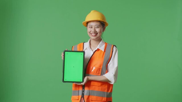 Asian Female Engineer With Safety Helmet Smiling And Showing Green Screen Tablet To The Camera While Standing In The Green Screen Background Studio

