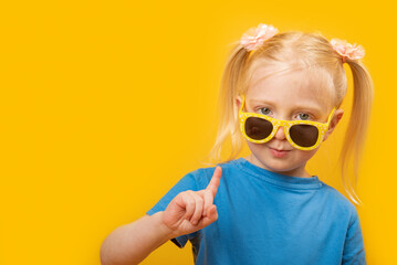 Portrait of little blonde girl with two tails in sunglasses showing finger, yellow background. Copy space, mock up
