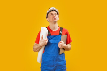 Man engineer or builder in blue work suit wears safety helmet and holds drawings and documents looking up. Isolated on yellow background.