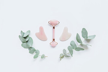Facial massage kit for home spa. Face roller  and gua sha massager made from rose quartz on white background with eucalyptus branches. Natural treatment concept. Flat lay. Top view