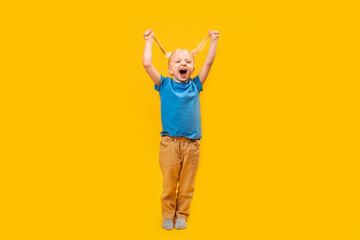 Full-length portrait of laughing little girl with two ponytails. Cheerful preschooler wears blue T-shirt on yellow background.