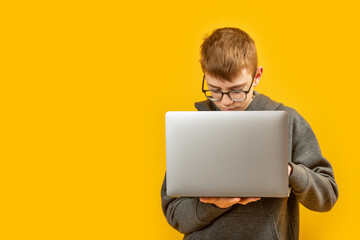 Boy with glasses holds laptop isolated on yellow background. Remote online learning. Programming for children. Copy space.