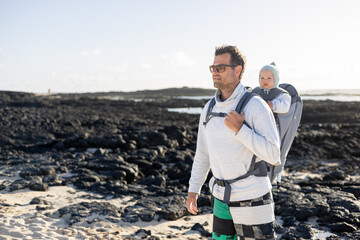 Young father carrying his infant baby boy son in backpack on black rock volcanic beach on Lanzarote island, Spain. Family travel and winter vacation concept - obrazy, fototapety, plakaty
