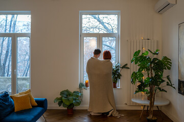 Family couple wife husband hugging looking at window standing in living room at home covering with knitted plaid, rear view. Affectionate man and woman in love cuddling together in cozy interior. - obrazy, fototapety, plakaty