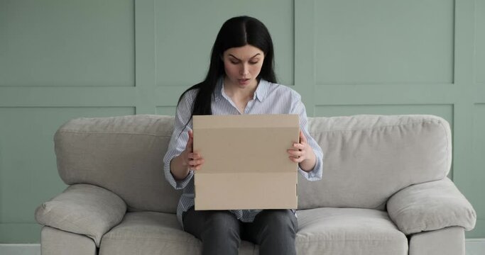 Caucasian woman receiving and eagerly opening a package, but becoming disappointed after seeing its contents. She closes the box and sits on the couch looking upset.