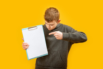 Brunette boy holds blank sheet of paper or documents and points his finger, isolated at them on yellow background. Copy space. Mock up.