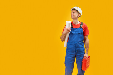 Handsome smiling man engineer worker or architect with helmet holds documents blueprints and tool box. Isolated on yellow background. Copy space.