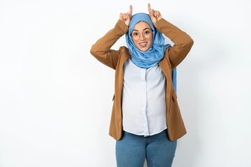 Funny Young beautiful pregnant muslim woman wearing hijab in white studio shows horns, fingers on head gesture, posing silly and cute