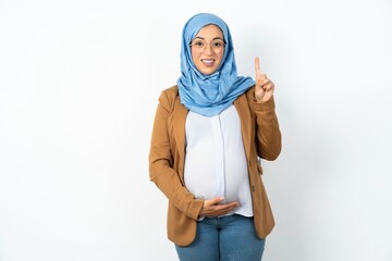 Young beautiful pregnant muslim woman wearing hijab in white studio showing and pointing up with finger number one while smiling confident and happy.