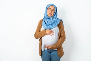 Displeased upset Young beautiful pregnant muslim woman wearing hijab over white wall frowns face as...