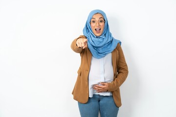 Excited positive Young beautiful pregnant muslim woman wearing hijab over white background points index finger directly at you, sees something very funny. Wow, amazing