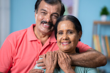 Obraz premium Close up head shot of happy indian senior couple embracing each other by looking at camera at home - concept of relationship, family bonding and relaxation