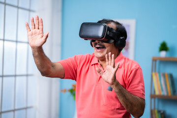 Happy indian senior man with VR or virtual reality headset experiencing metaverse at home - concept of imagination, futuristic and technology
