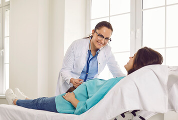 Friendly gynecologist doctor examining heartbeat in the abdomen of pregnant young woman lying on the couch in medical clinic. Obstetrician doctor checking her patient expecting a baby.