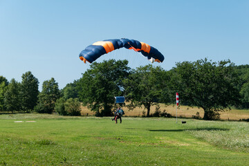Paraquedista a fazer o pouso junto ao solo num campo verde rodeado por floresta