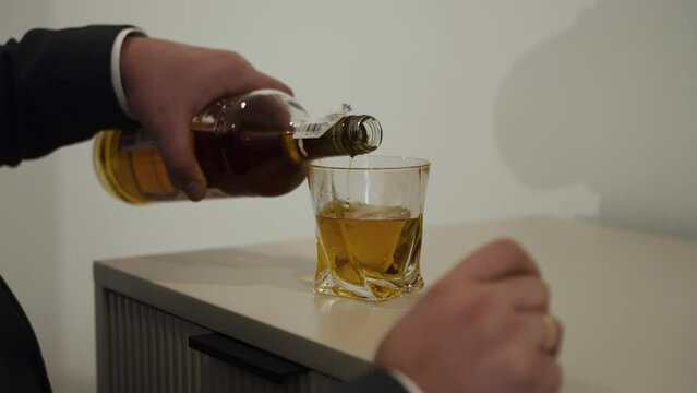 A person pours amber-colored liquid, likely whiskey, into a clear glass. The setting suggests a tidy, minimalist space with a focus on the action of pouring. The image captures the elegance 
