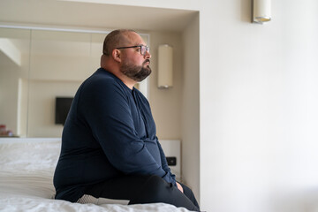 Sad fat lonely man sitting on bed in hotel and looking out window, thinking about life problems, no strength change, side view overweight depressed man no friends and mental disorders - obrazy, fototapety, plakaty