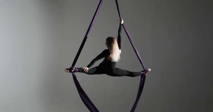 talented Caucasian dancer on the aerial silks. Against a dark studio background, her long hair cascading down. Clinging to the silk ribbons, she effortlessly executes mesmerizing wraps, spins.