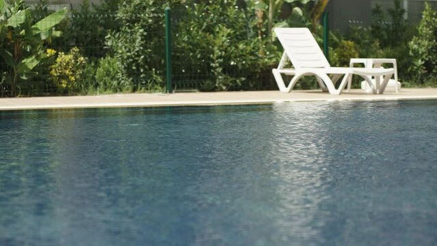 A view of the blue water of the pool and a white lounge chair against the backdrop of bushes and a fence.