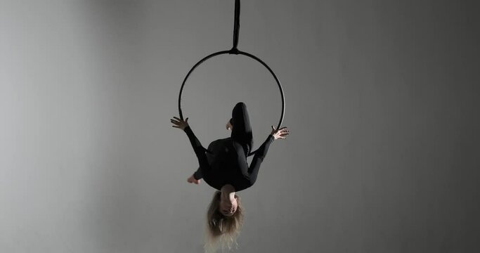 On a darkened stage, a caucasian dancer with her aerial hoop performance. Dressed in a black jumpsuit and barefoot, she moves with confidence, utilizing the hoop as an extension of body.
