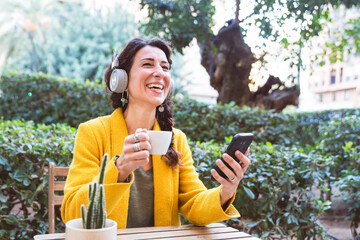 Adult woman with headphones holds a mobile phone and laughs on a park terrace