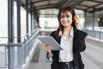 Attractive young woman in black suit holding tablet wearing headphone smiling looking a camera standing at public places. Lifestyle people technology concept. Female student outside university.