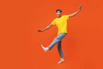 Fototapeta na wymiar Full body young man wear pyjamas jam sleep eye mask rest relax at home look camera with outstretched hands raise up leg isolated on plain orange background studio portrait Good mood night nap concept