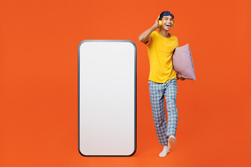 Full body young man wear pyjamas jam sleep eye mask relax at home big blank screen area mobile cell phone smartphone listen music in headphones isolated on plain orange background. Night nap concept.
