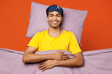 Calm young happy man wear pyjamas jam sleep eye mask rest relax at home lies wrap covered under blanket duvet close eyes isolated on plain orange color background studio. Good mood night nap concept.