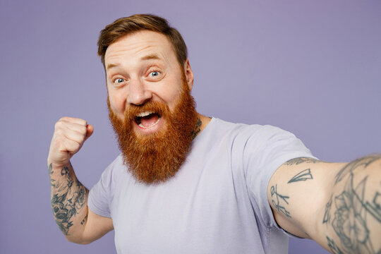 Close up young redhead bearded man wear violet t-shirt casual clothes doing selfie shot pov on mobile cell phone do winner gesture isolated on plain pastel light purple background Lifestyle concept