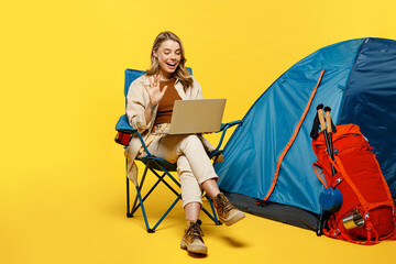 Full body young IT woman sit near bag with stuff tent hold use work on laptop pc computer wave hand isolated on plain yellow background Tourist walk on spare time Hiking trek rest travel trip concept