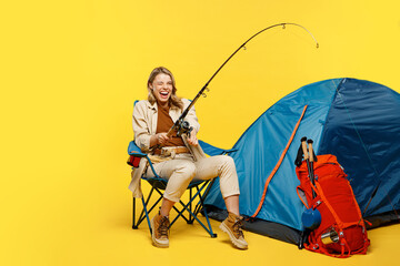Full body happy young woman sit near bag with stuff tent hold fishing rod isolated on plain yellow...
