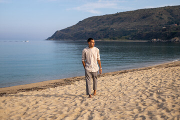 young caucasian man in collarless shirt and dress pants walking on the beach