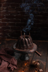 Homemade chocolate bundt cake on a wooden stand, napkin, a cup of fresh made coffee on wooden rustic table. Brick wall background. Still life. Dark and moody image. - 611049426