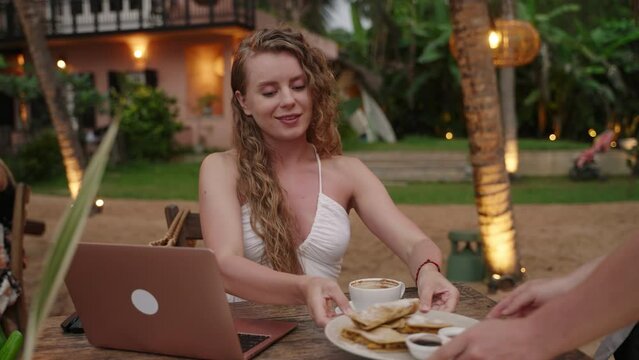 Caucasian woman with laptop in outdoor cafe at resort drinks coffee and starts working. Female freelancer typing on computer at tropical location gets coffee from waiter smiling and continues her work