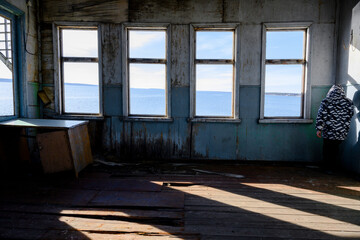 A boy in the corner of a room of an abandoned wooden house with empty windows without glass and a view of the river