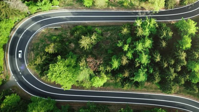 Aerial view from above down a scenic mountain road in a coniferous forest. Car drives along a twisting road in alpine terrain. Scenic serpentine uphill path in the light of sunset on a sunny day. 4k