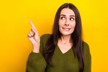 Photo of doubtful uncertain woman dressed khaki sweater biting lip pointing finger looking empty space isolated yellow color background