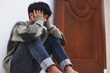 Depressed man sitting on the floor with head in hands and leaning against a wall. 