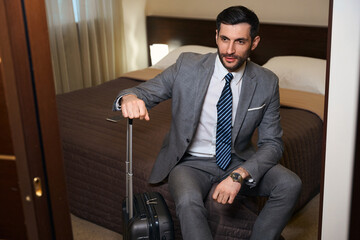 Traveling man with a travel suitcase sits on large bed