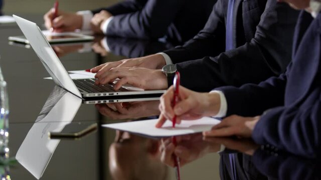Meeting participants write down important points on laptop or sheets of paper quickly. Persons sit near black long office table crop body