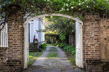 Obraz premium Looking down an alley through an open gate to the two-track gravel road along the side of a home with some sunlight.