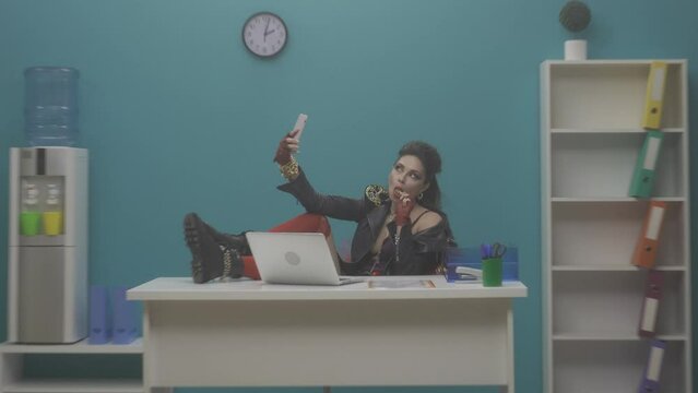 A woman freak, being at the workplace, takes a selfie on a smartphone, eats a lollipop. A woman sits in the office with her feet on the table. Content. Feminism. Advertising. HDR BT2020 HLG Material
