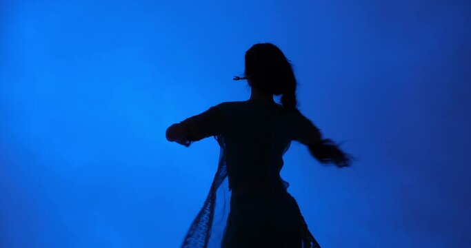 Young Indian woman performs a traditional Indian dance in a studio against a blue background. She wears a beautiful sari, along with the jewelry commonly worn by Indian dancers.