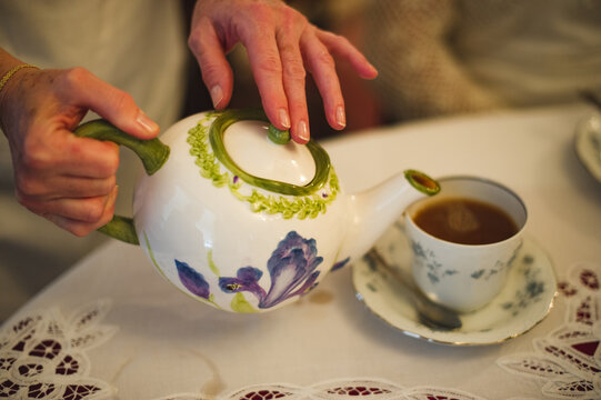 A woman's hands pour tea from an antique pot into a glass on a white tablecloth